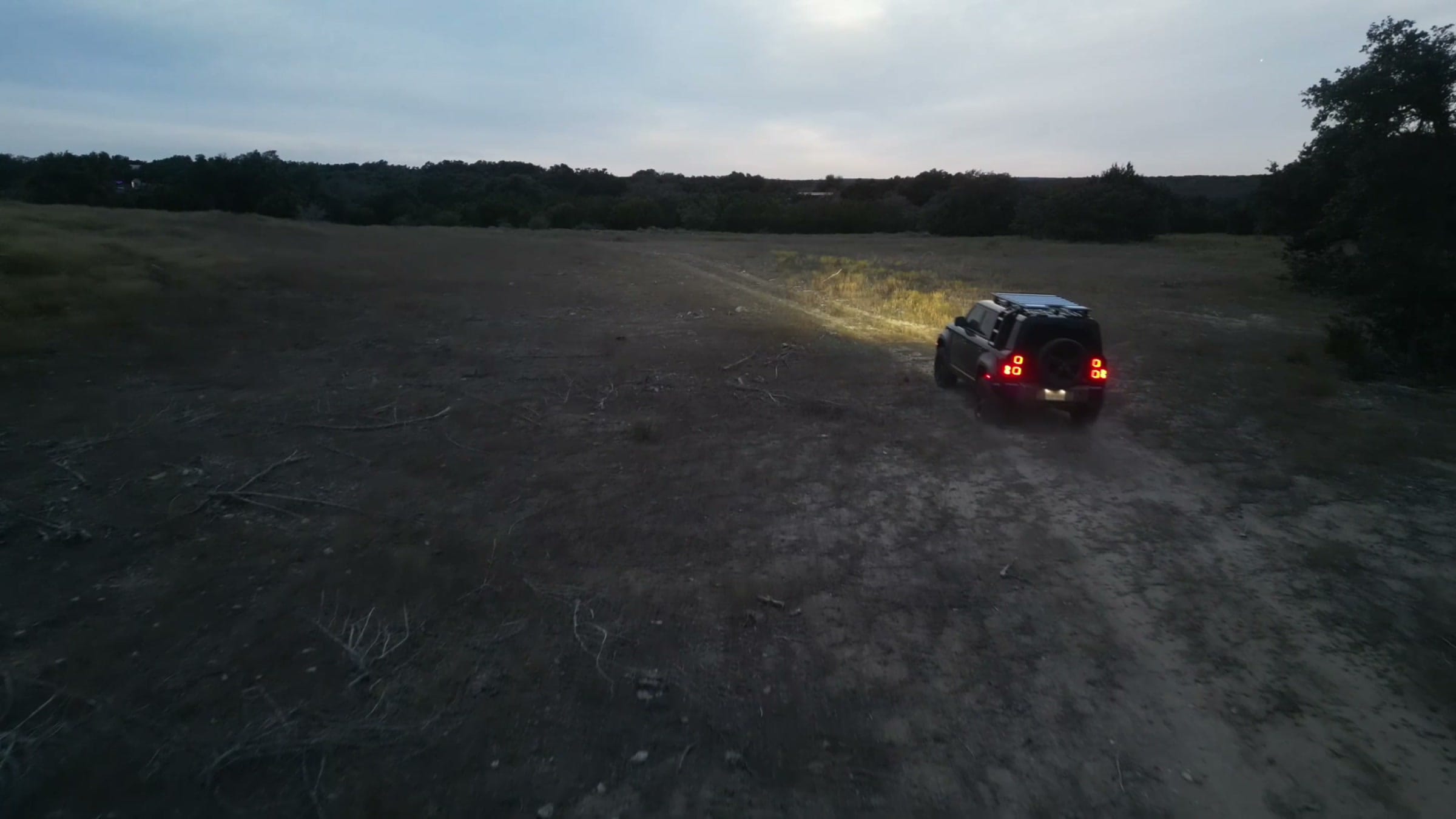 Drone view of an SUV crossing open pasture at dusk with its headlights on